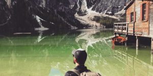 man in brown jacket sitting on brown rock near body of water during daytime